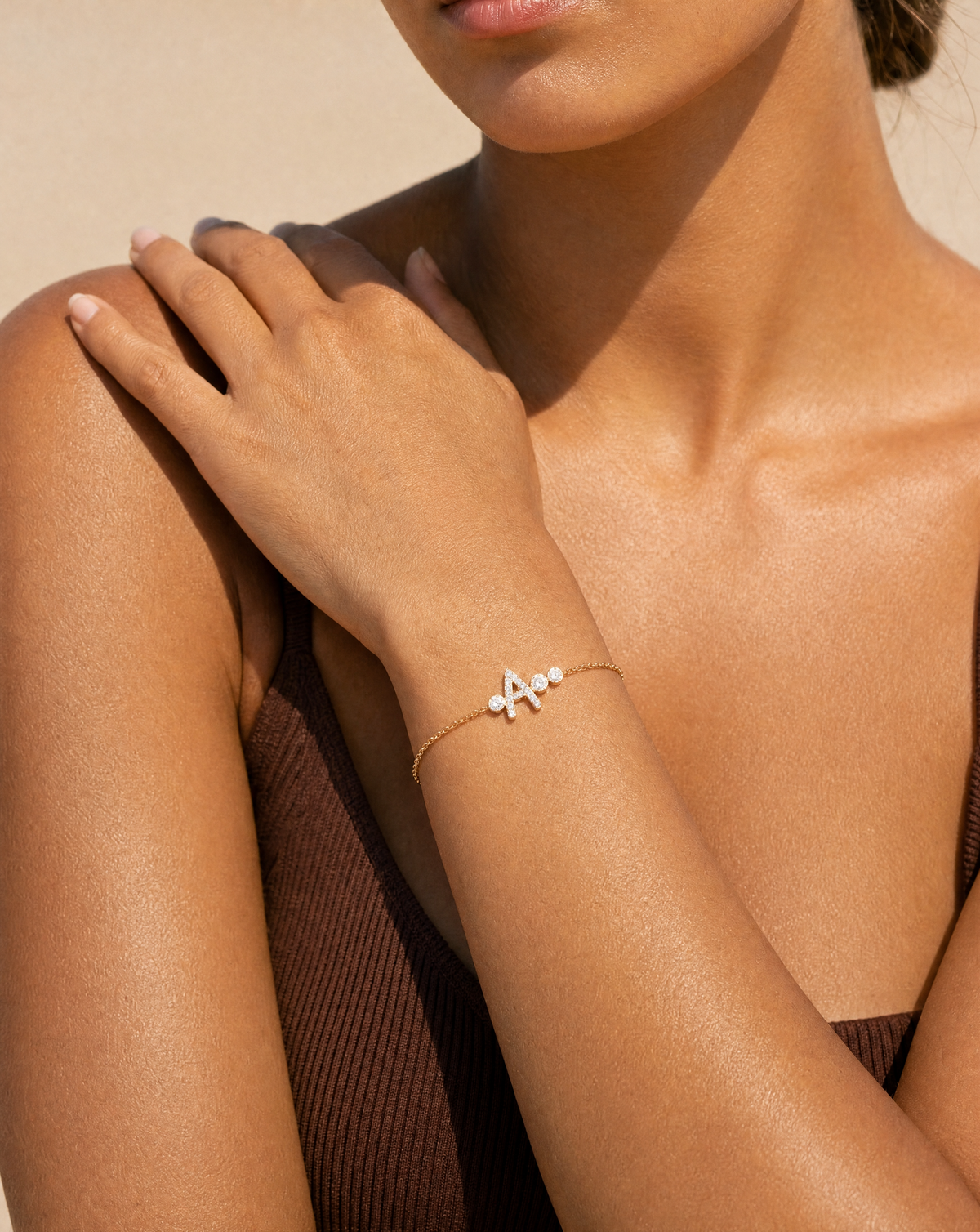 A close-up lifestyle shot of a model wearing a delicate gold chain bracelet on her wrist. The bracelet features a small gold letter 'A' accented with tiny white dots, set against a warm beige background.