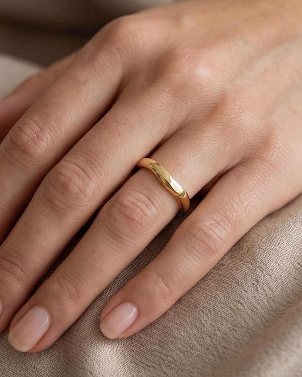A close-up of a hand wearing a thin, polished gold band ring on the ring finger, resting on a soft beige fabric surface.